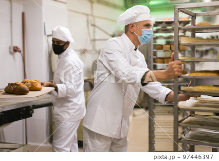 man in chefs uniform rolling trolley with bread in bakery during epidemic 97446397