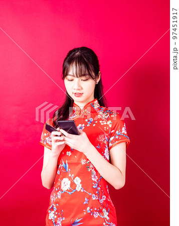 赤い背景にスマートフォンを使って、伝統的なチャイナドレスを着たアジアの若い女性のポートレート。 赤い背景にスマートフォンを使って、伝統的なチャイナドレスを着たアジアの若い女性のポートレート。 97451917