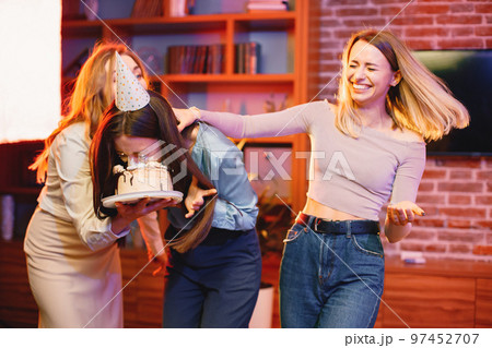 Young woman dips face in white cake with cream. Three girls having fun together. Birthday face cake smashing tradition concept. Young woman dips face in white cake with cream. Three girls having fun together. Birthday face cake smashing tradition concept. 97452707