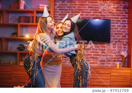 Young friends standing in indoors in evening party red light. Girls having fun under a confetti. Everybody wearing party cones. 97452709