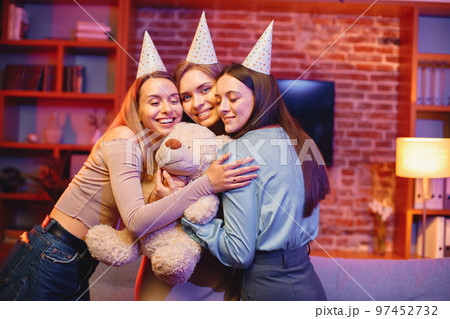 Young friends standing in indoors in evening party red light. Everybody wearing party cones. Girl holding a bear toy gift. 97452732