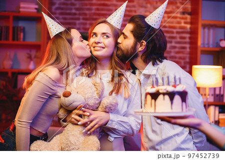 Young friends standing in indoors in evening party red light. Everybody wearing party cones. Boy and girls kissing girl in a middle in a cheek. 97452739