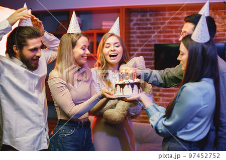 Young friends standing in indoors in evening party red light. Everybody wearing party cones. Boys and girls holding a birthday cake and light a candles. 97452752