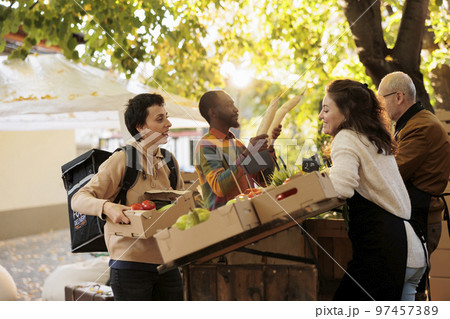 Cheerful courier and local stand owner talking about natural products, wokring on delivering fresh food order to clients. Greengrocer giving box with fruits and veggies to delivery woman. Cheerful courier and local stand owner talking about natural products, wokring on delivering fresh food order to clients. Greengrocer giving box with fruits and veggies to delivery woman. 97457389
