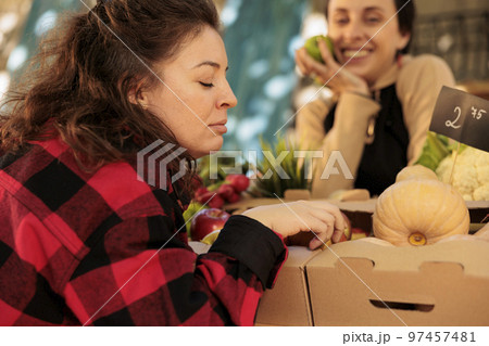 Young woman choosing fruits and vegetables while shopping at local farmers market. Female customer smelling and touching colorful apples while standing at fresh organic produce stand Young woman choosing fruits and vegetables while shopping at local farmers market. Female customer smelling and touching colorful apples while standing at fresh organic produce stand 97457481