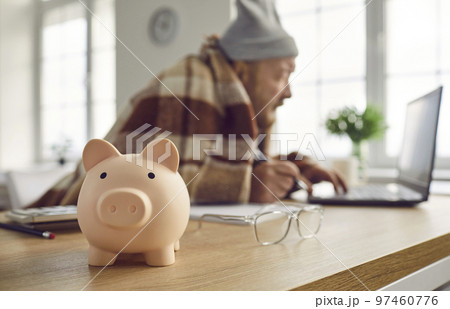 Close up of piggy bank on table with senior retired man using laptop computer in background Close up of piggy bank on table with senior retired man using laptop computer in background 97460776