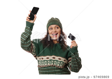Attractive joyful brunette rejoicing, smiling, looking at a copy space on white background, posing with modern smartphone and credit card in her hands. Free space for your advertising and mobile apps 97469869
