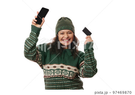 Cheeful dark-haired woman in green warm clothes posing with smartphone and credit card in her hands, smiling a beautiful toothy smile, looking at camera, isolated over white background. Copy ad space 97469870