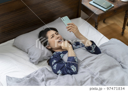 ベッドに寝ながらスマホを見て顔をしかめるアラフィフの女性 97471834
