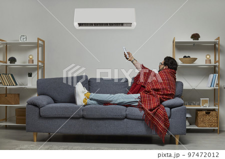 Young man wrapped in blanket holding air conditioner remote control 97472012