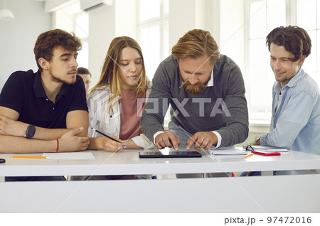 Male teacher in class helps group of highschool students working on digital tablet. 97472016