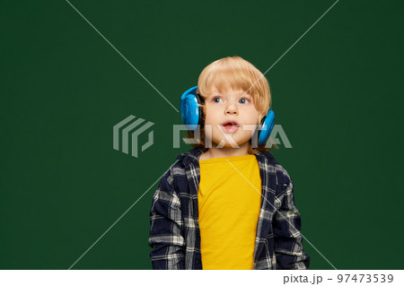 Portrait of little boy, child posing in headphones, listening to fairy tales over green studio background. Concept of childhood, emotions 97473539