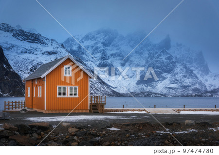 Orange rorbu on sea coast and snow covered mountain in fog Orange rorbu on sea coast and snow covered mountain in fog 97474780
