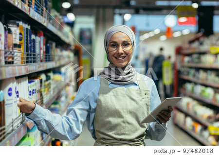Portrait of a female saleswoman in a hijab, a salesperson in a household chemicals department is smiling and looking at the camera, holding a laptop tablet computer in her hands. Portrait of a female saleswoman in a hijab, a salesperson in a household chemicals department is smiling and looking at the camera, holding a laptop tablet computer in her hands. 97482018