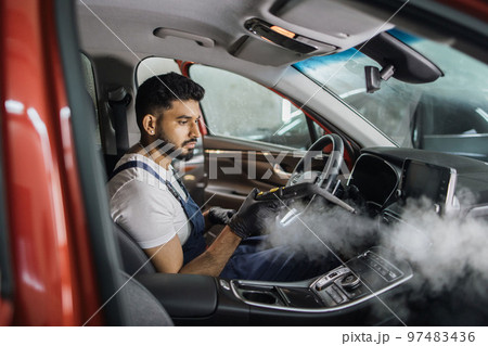 Handsome bearded man in blue uniform overalls cleaning interior of the car with hot steam cleaner. 97483436
