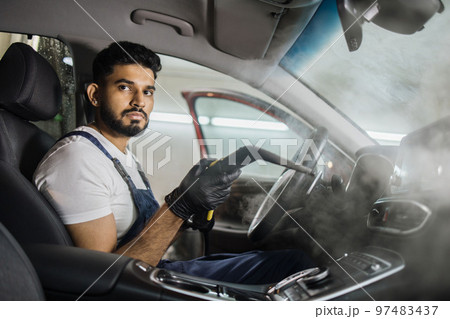 Handsome man in overalls, worker of car wash center, cleaning car interior with hot steam cleaner. 97483437