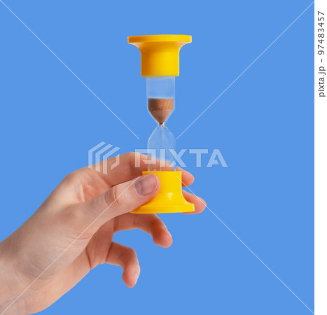 Yellow hourglass with running sand in female hand over blue background 97483457