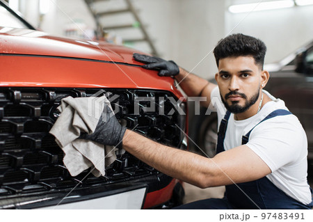 Young bearded male professional car wash worker, holding the gray microfiber and wiping grille 97483491