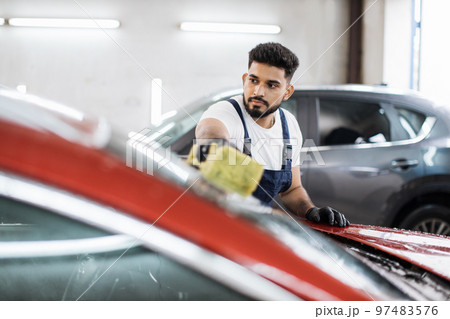Young bearded man, car wash worker, wearing t-shirt and overalls cleaning the windshield of car Young bearded man, car wash worker, wearing t-shirt and overalls cleaning the windshield of car 97483576