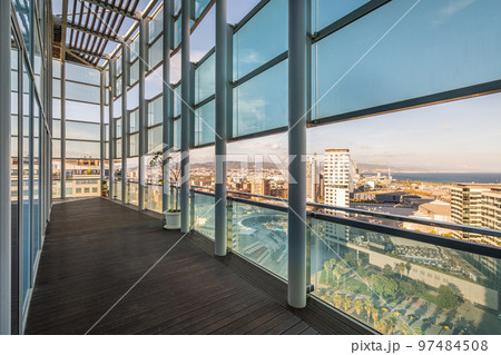 Long spacious terrace in high-rise building with fence and roof made of metal beams of strong impact-resistant glass protected from direct sunlight. Floor of terrace is covered with wooden parquet. 97484508