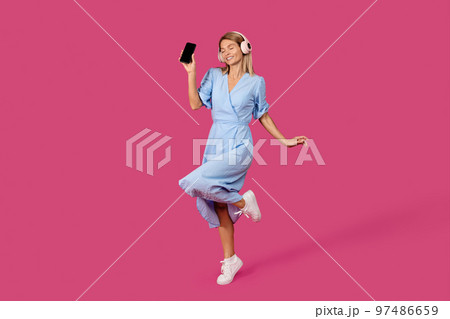 Cheerful mature caucasian female blonde in dress and wireless headphones shows smartphone with empty screen Cheerful mature caucasian female blonde in dress and wireless headphones shows smartphone with empty screen 97486659