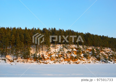 Winter landscape with the moon in the sky during the day. The river is covered with snow. Pine trees grow on red stones. Nature of Eastern Siberia. Winter landscape with the moon in the sky during the day. The river is covered with snow. Pine trees grow on red stones. Nature of Eastern Siberia. 97501733