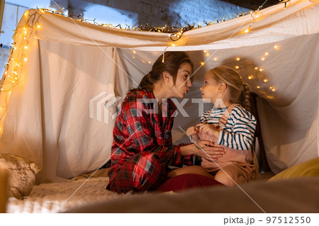 Happy family. Mother and daughter in pajamas sitting inside self-made hut, tent in room in the evening. Cozy time Happy family. Mother and daughter in pajamas sitting inside self-made hut, tent in room in the evening. Cozy time 97512550