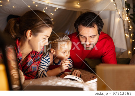 Happy family, mother, father and daughter in pajamas lying inside self-made hut, tent in room in the evening and reading stories Happy family, mother, father and daughter in pajamas lying inside self-made hut, tent in room in the evening and reading stories 97512565