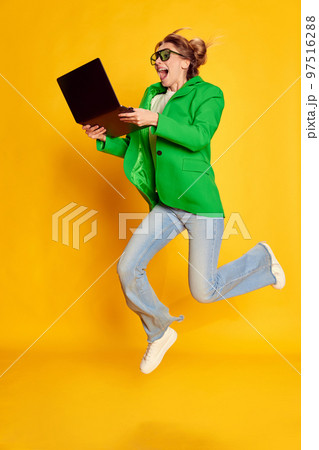 Portrait of young girl in bright green jacket posing with laptop in a jump over yellow studio background. Successful project 97516288