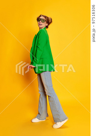 Portrait of young girl in bright green jacket posing over yellow studio background. Stylish, fashionable youth 97516306