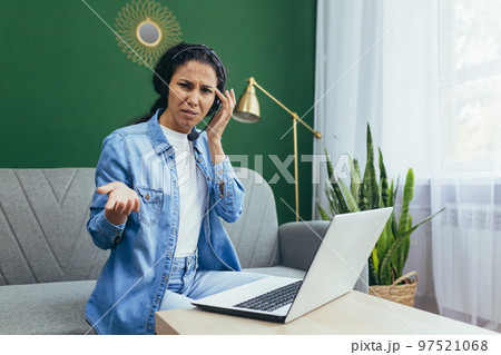 Portrait of frustrated and upset woman at home, Hispanic woman working remotely looking at laptop at work and video call headset sitting on sofa in living room. 97521068