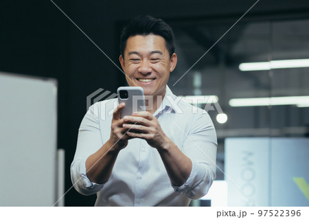 A young smiling Asian man is standing in the office and using the phone. Plays online games, types messages, reads good news. A young smiling Asian man is standing in the office and using the phone. Plays online games, types messages, reads good news. 97522396