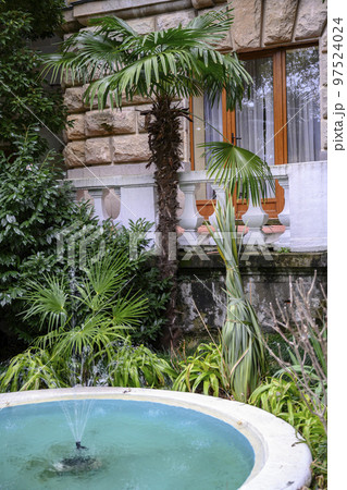 Fragment of a building with a balcony in a garden with a palm tree and a fountain Fragment of a building with a balcony in a garden with a palm tree and a fountain 97524024