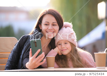 Young mother and her child daughter taking picture with phone selfie camera sitting at street cafe with hot drinks on sunny fall day. Social media presence in everyday life concept Young mother and her child daughter taking picture with phone selfie camera sitting at street cafe with hot drinks on sunny fall day. Social media presence in everyday life concept 97525434