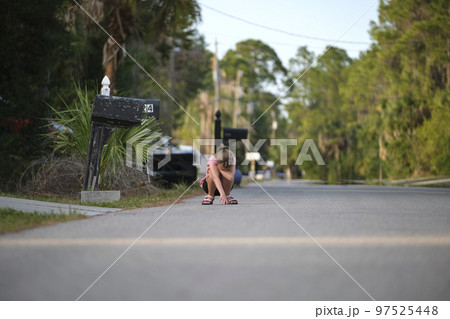 Young moody child girl squatting in the middle of the road covering face with hands on sunny day Young moody child girl squatting in the middle of the road covering face with hands on sunny day 97525448
