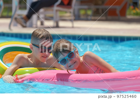Young joyful children boy and girl having fun swimming on inflatable air mattress in swimming pool with blue water on warm summer day on tropical vacations. Summertime activities concept Young joyful children boy and girl having fun swimming on inflatable air mattress in swimming pool with blue water on warm summer day on tropical vacations. Summertime activities concept 97525460
