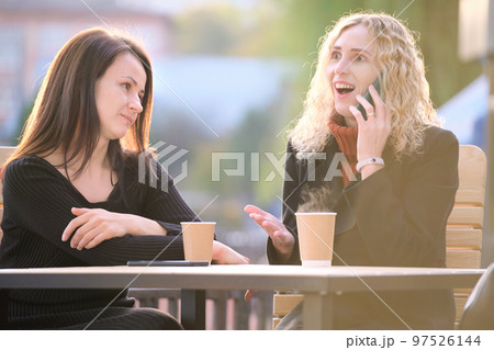 Unhappy young woman waiting angrily while her friend is talking happily on sellphone with someone else and ignoring her. Friendship problems concept 97526144