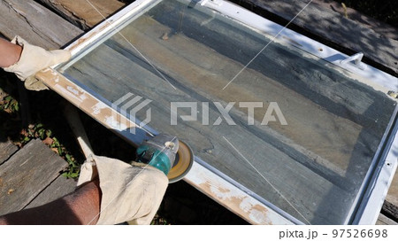sanding a white-painted wooden frame, scraping off old paint and smoothing the surface of the wood with a grinder in the hands of a carpenter in canvas mittens in a street workshop sanding a white-painted wooden frame, scraping off old paint and smoothing the surface of the wood with a grinder in the hands of a carpenter in canvas mittens in a street workshop 97526698