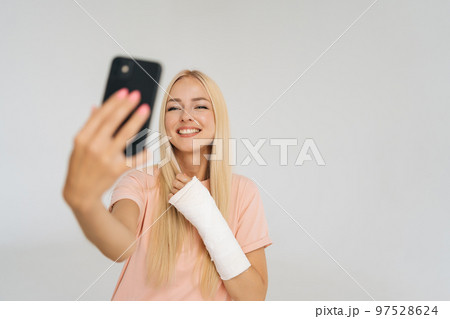 Studio portrait of joyful female blogger with broken arm wrapped in plaster bandage talking smartphone making video call, taking selfie picture, standing on white isolated background. Studio portrait of joyful female blogger with broken arm wrapped in plaster bandage talking smartphone making video call, taking selfie picture, standing on white isolated background. 97528624