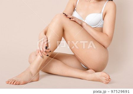 Cropped shot of half naked young woman wears white lingerie, shows slender legs, has healthy smooth skin, crossed legs, sits on floor against beige background, has perfect figure, poses bare foot. Cropped shot of half naked young woman wears white lingerie, shows slender legs, has healthy smooth skin, crossed legs, sits on floor against beige background, has perfect figure, poses bare foot. 97529691