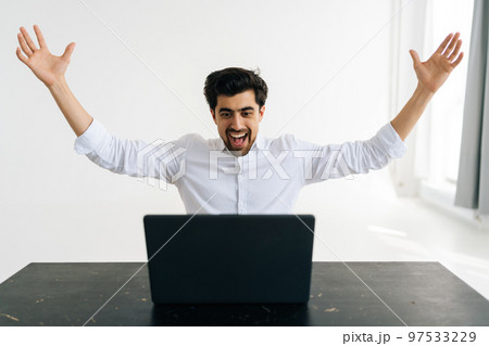 Studio portrait of cheerful excited male office worker screaming eureka raising hands up, solving hard task, getting access, successfully completing work sitting at desk in room with light interior. 97533229