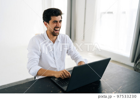 Studio portrait of happy excited young man in shirt using working on laptop computer smiling looking away sitting at table in room with light interior. Cheerful male freelancer at remote home office. 97533230