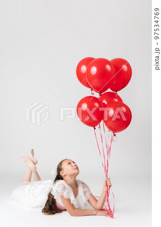 Beautiful happy girl on birthday holiday party, lying down and holding large bunch of red balloons in hand, looking up at balls. Studio shot of little girl on white background. Part of photo series. 97534769