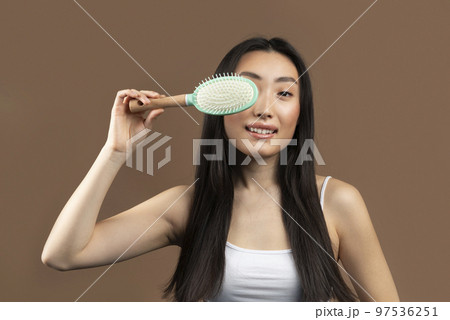 Hair care concept. Excited korean woman with long hair covering eye with hair brush and smiling over brown background 97536251