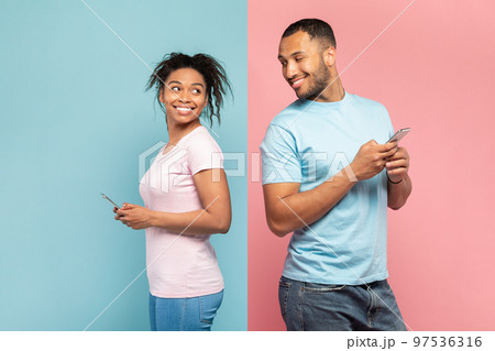 Curious black man using smartphone, looking and peeking at his girlfriend's phone, standing on pink and blue background Curious black man using smartphone, looking and peeking at his girlfriend's phone, standing on pink and blue background 97536316