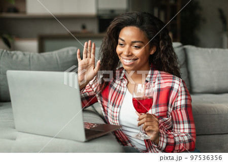 Glad millennial black lady waving hand, say hello greeting in computer camera, hold glass of wine in living room Glad millennial black lady waving hand, say hello greeting in computer camera, hold glass of wine in living room 97536356