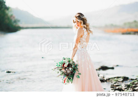 Beautiful brunette bride in elegant white dress holding bouquet posing near river Beautiful brunette bride in elegant white dress holding bouquet posing near river 97540713
