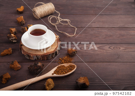 Chaga mushroom coffee drink in a white cup on a wooden stand, scattered pieces of chaga and a spoon with chaga powder on a wooden background. Copy spaes. 97542102