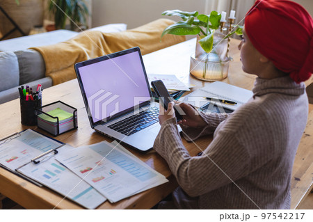 Biracial woman wearing hijab, sitting at table in living room and using laptop with copy space Biracial woman wearing hijab, sitting at table in living room and using laptop with copy space 97542217