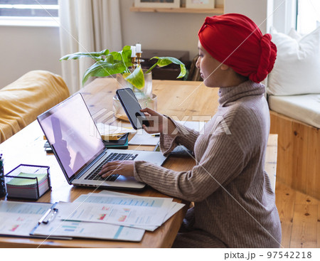 Biracial woman wearing hijab, sitting at table in living room and using laptop with copy space Biracial woman wearing hijab, sitting at table in living room and using laptop with copy space 97542218